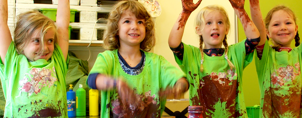 Kinder im Kindergarten bei einem Kunstprojekt, gefördert durch Fröbel-Fördermitgliedschaften. Sie malen mit Farbe und tragen grüne Malkittel, die mit bunten Farbe bekleckst sind. Sie recken die Hände in die Höhe und lachen in die Kamera. 