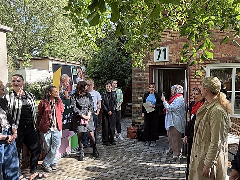Eine Gruppe von Menschen steht vor dem Eingang eines Backsteingebäudes, das das Familienzentrum Treptow 71 ist. Das Gebäude hat eine weiße Nummer „71“ über der braunen Eingangstür. Grüne Bäume und Sträucher umrahmen das Bild, und helles Sonnenlicht fällt durch die Blätter.